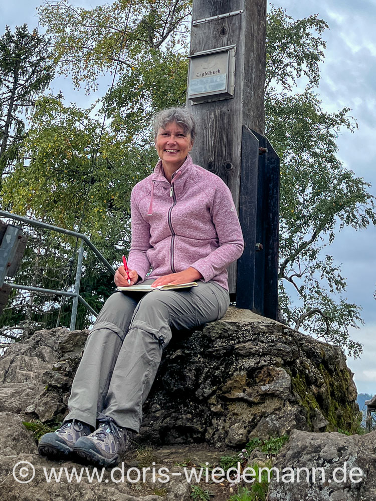 20250823-unbenannt-IMG_0437 Doris Wiedemann sits at the summit cross of the Hennenkobel in the Bavarian Forest and writes an entry in the summit book