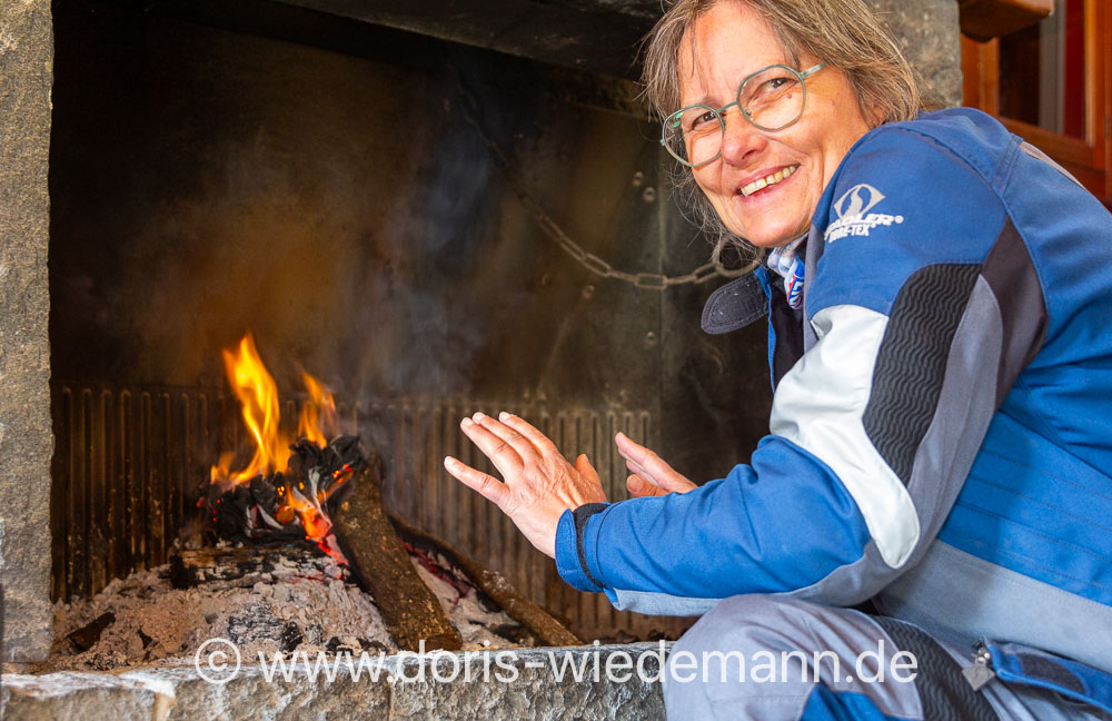Doris Wiedemann sitzt in Motorradkleidung vor einem offenen Kamin und wärmt sich die Hände am Feuer